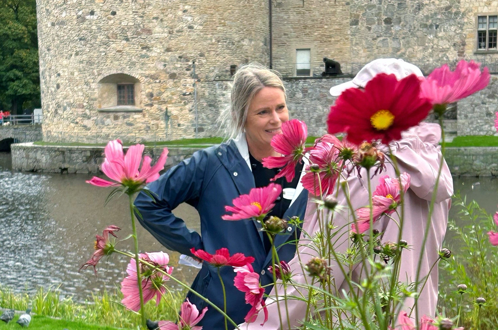 En leende kvinna i regnrock samtalar med en äldre kvinna med rollator. I bakgrunden Örebro slott, i förgrunden rosa, höga blommor.