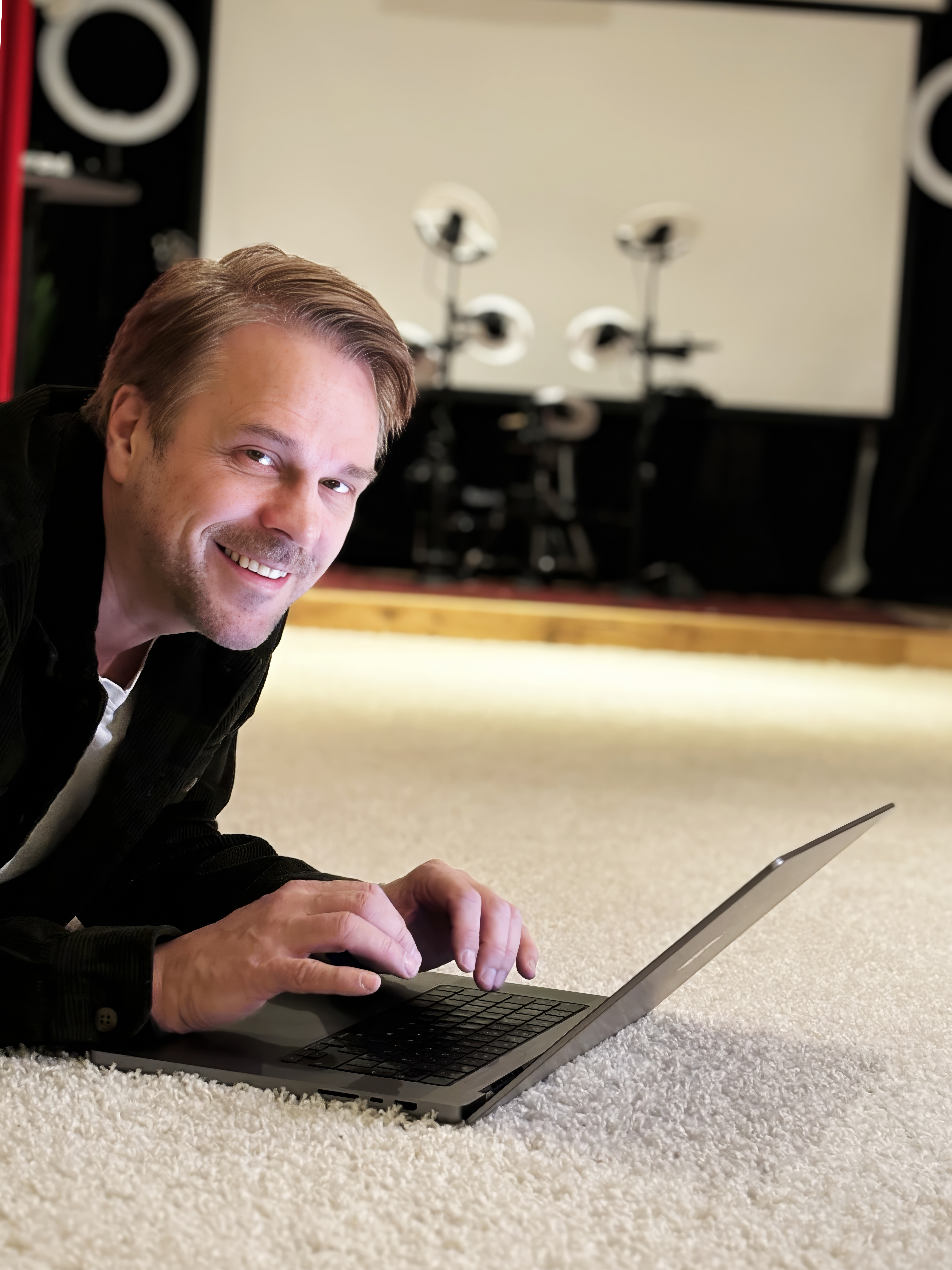 En man ligger på golvet inuti ett hus och ler medan han snappar på sin laptop.