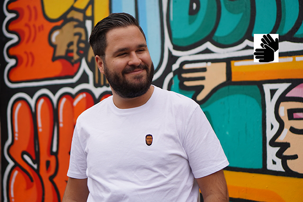 En skäggig man med bakåtkammat hår står framför en graffittimålning. Ikon för teckenspråkstolkning