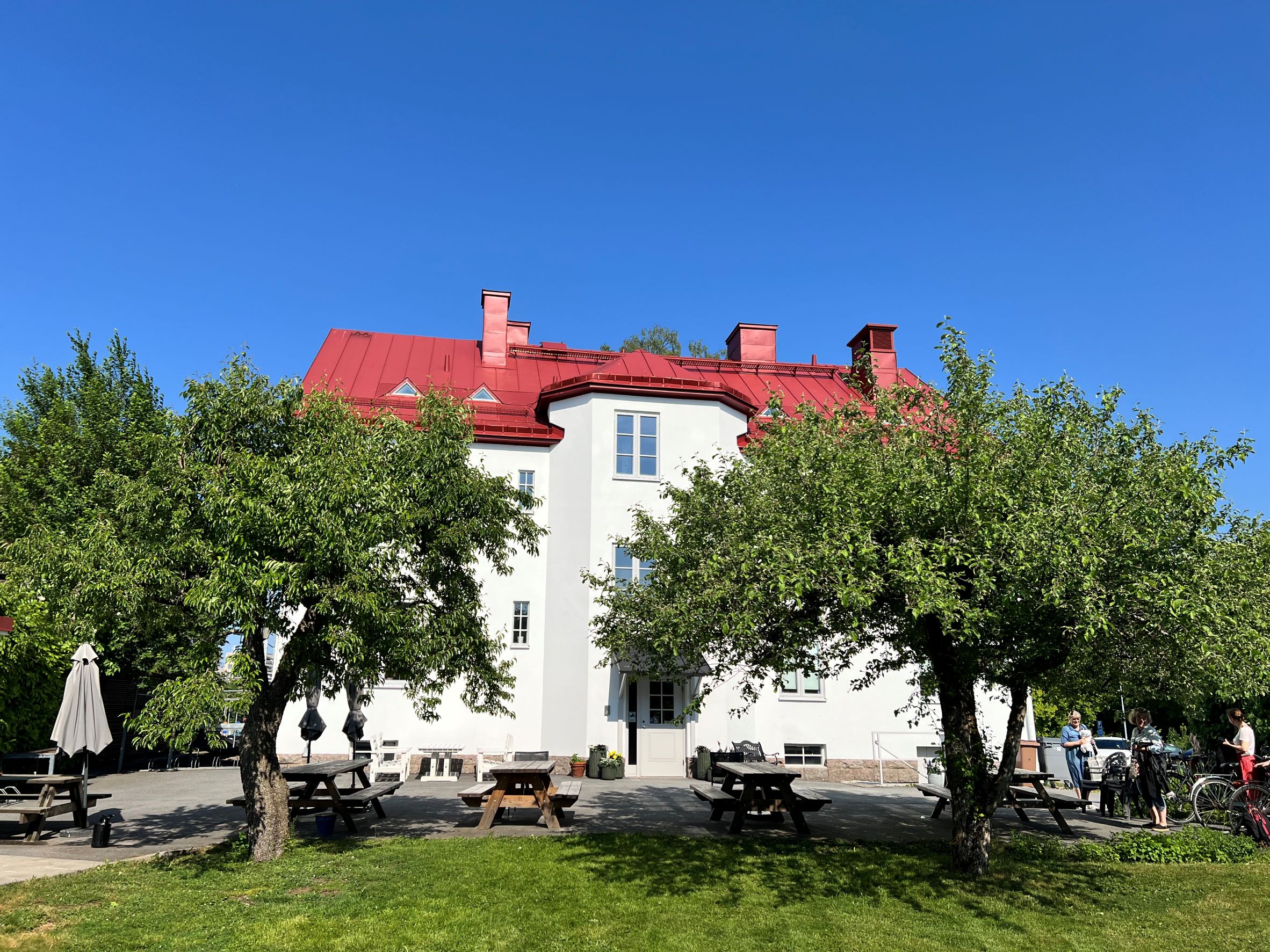Två gröna träd står framför ett stort vitt hus med rött tak. Himlen är klarblå.