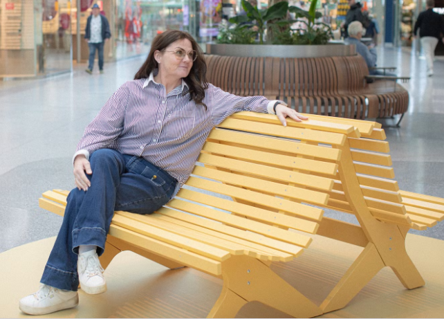 En person sitter på en gul bänk. Det är en bänk med sittyta åt två håll. 