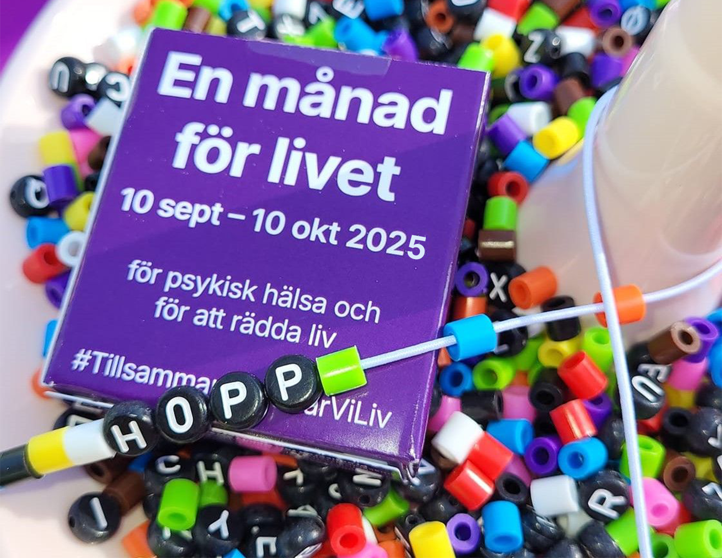 I ett av av pärlor i olika färger ligger en lila tablettask med information om En månad för livet 2025. Ovanpå asken ligger ett pärlband med pärlor på där det står "Hopp".