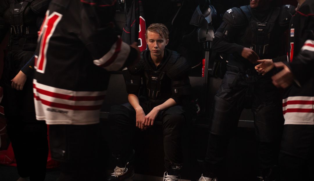 En ung kille med skridskor och hockeyskydd sitter på en bänk i omklädningsrummet. Runt honom står andra i spelartröjor.